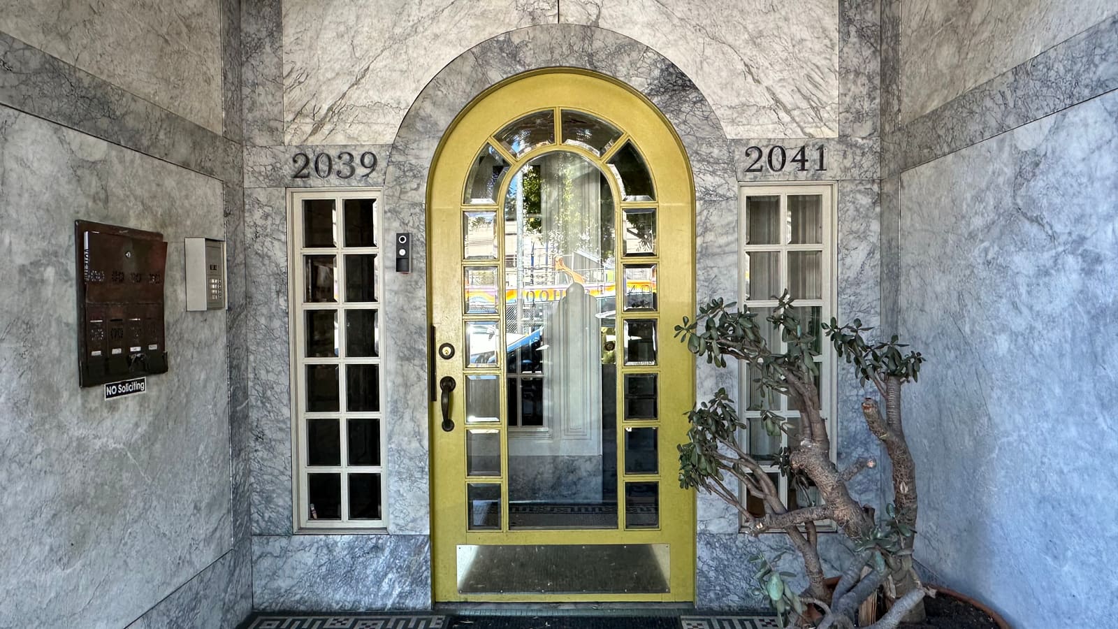 A yellow arched front door set between the street numbers 2039 and 2041, with a shared bank of mailboxes and a buzzer panel on the marble wall beside it.