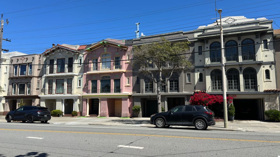 A row of Edwardian flats in muted pink, grey and tan along a wide Inner Richmond street.