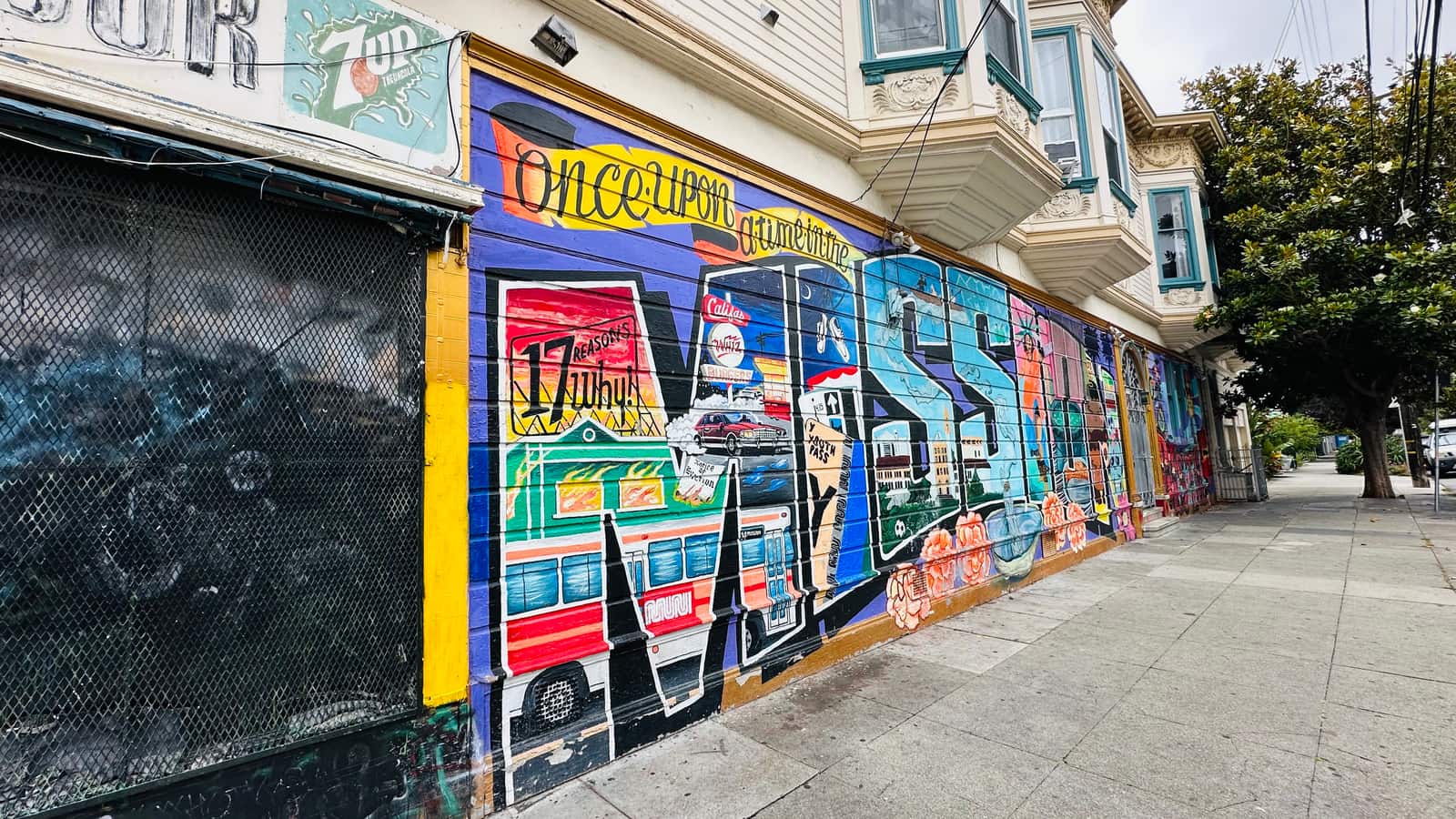 A full-wall mural in saturated colour along the flank of a Mission District Victorian, the block of houses rising behind it.