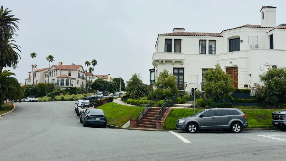 White Mediterranean-style houses with palms along a curved Sea Cliff street under overcast sky.