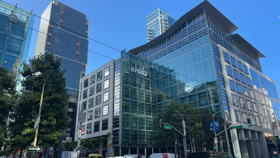 A glass-fronted office tower in San Francisco's Financial District seen from street level, with taller towers rising behind it.