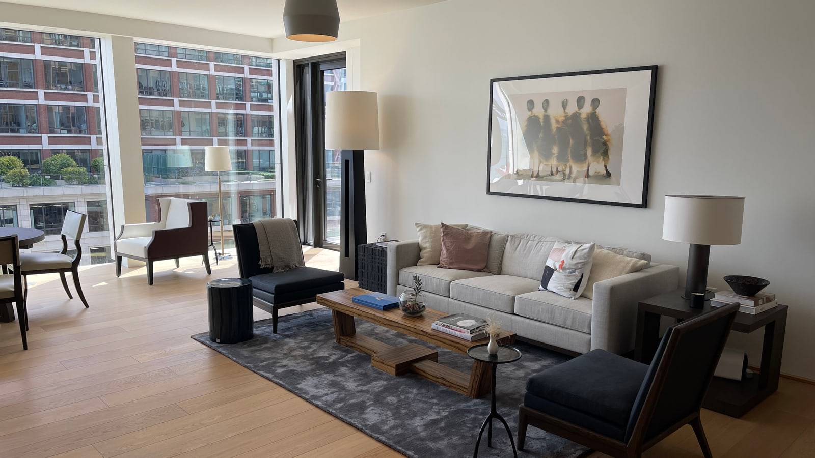 A staged condominium living room with pale sofas, framed artwork and floor-to-ceiling windows onto neighbouring buildings.