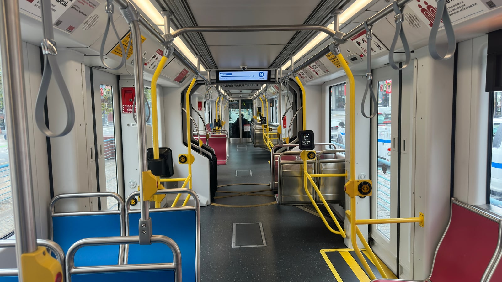 The interior of an empty Muni Metro light-rail car, yellow grab poles and hanging straps down the aisle.
