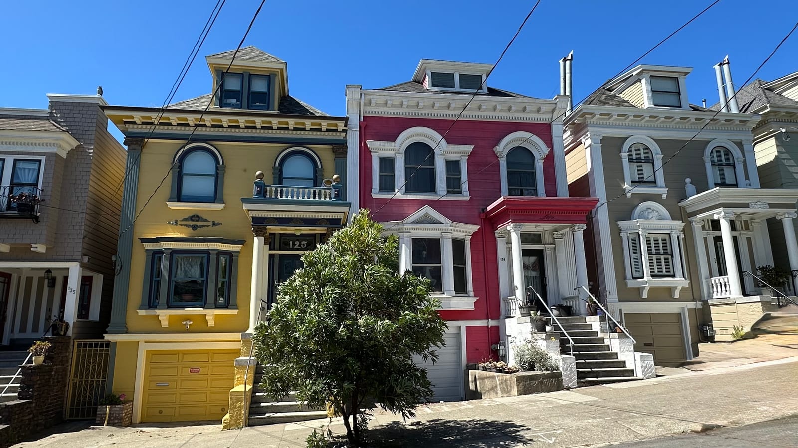 A row of Victorian houses in yellow, crimson and olive, their bay windows and painted trim in full sun on a sloping street.