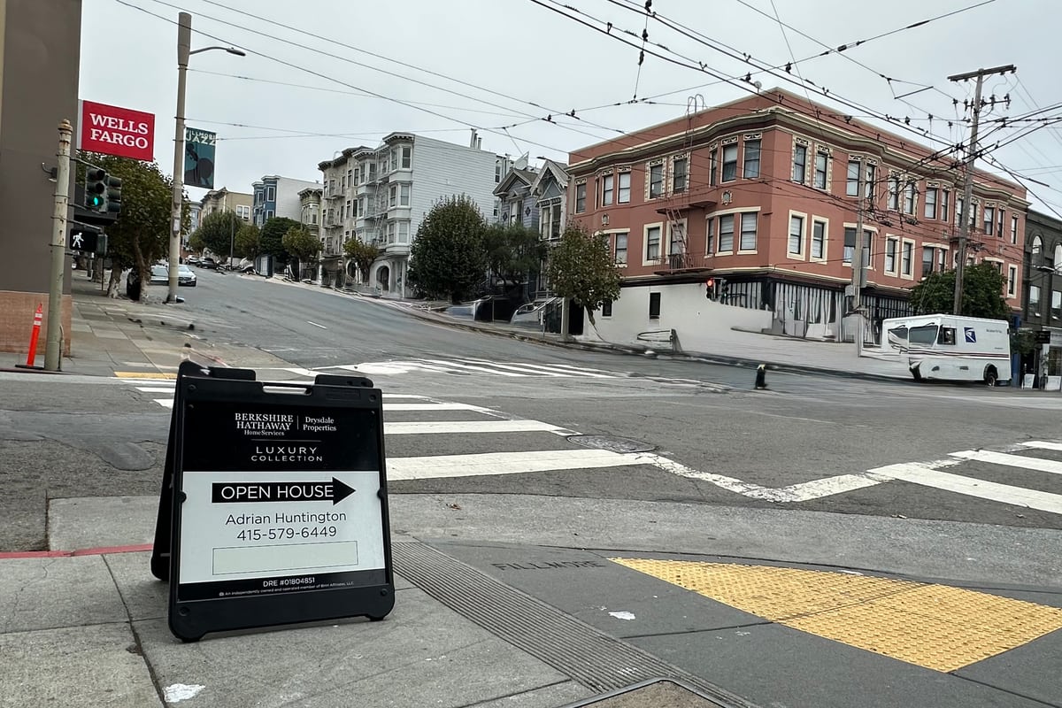An open-house A-frame sign on a Fillmore Street corner, with Victorian flats climbing the hill behind it on an overcast San Francisco morning.