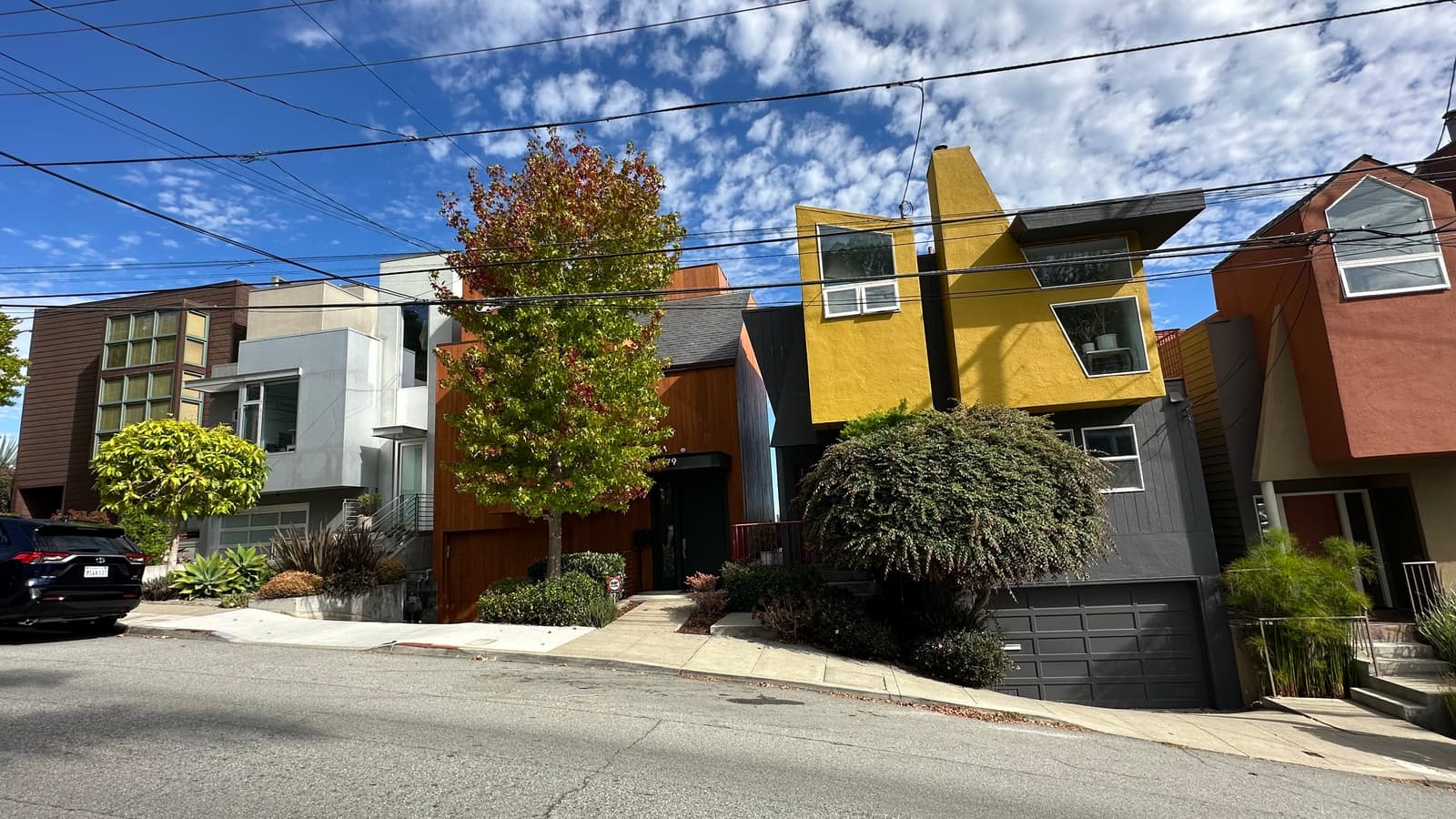 A row of contemporary houses stepping up a steep Bernal Heights block, cedar and white stucco beside a mustard-yellow facade and a maple turning red.
