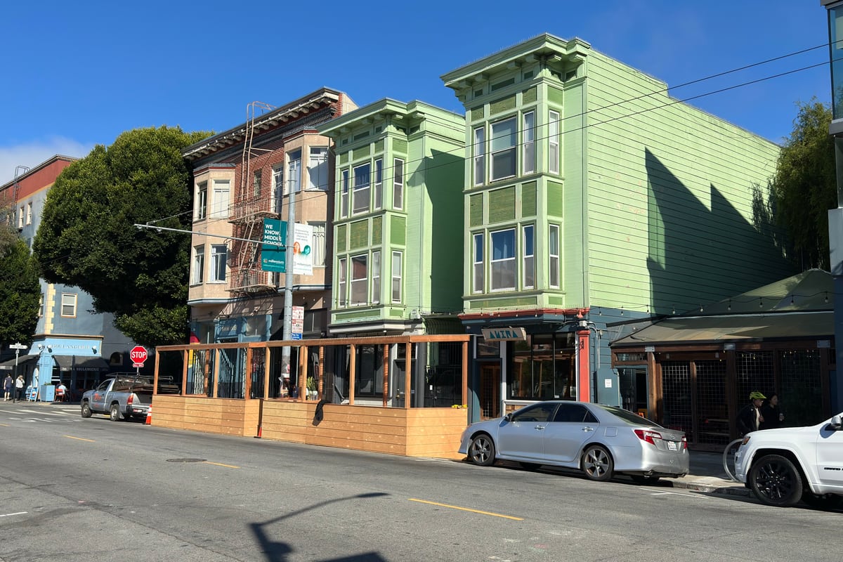 A pale green Victorian corner building above a restaurant parklet on a sunny Hayes Valley street in San Francisco.