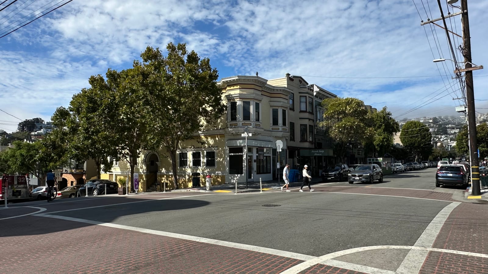 The corner of 24th and Sanchez Streets in Noe Valley, with a pale-yellow Victorian above ground-floor shops, mature street trees, and the 24th Street shopping strip running east toward the hills.