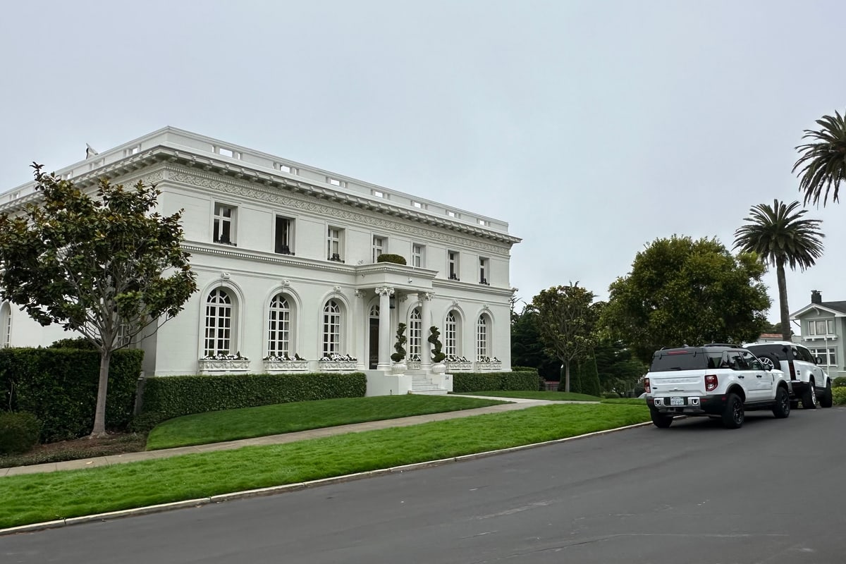 A white neoclassical mansion behind a clipped hedge on a quiet Presidio Heights street in San Francisco, palms rising beyond the roofline.